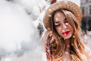 Wall Mural - Romantic girl with red lips posing with eyes closed near sparkle ball. Outdoor shot of pensive jocund woman in straw hat.