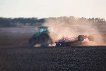 Tractor with a disc harrow system harrows the cultivated farm field, process of harrowing and preparing the soil, tractor seeding crops at field on sunset, agriculture concept, harrow machine at work