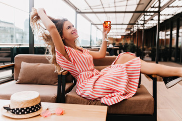 Ecstatic blonde girl playing with her hair while chilling in cafe. Inspired young woman in striped dress enjoying champagne in restaurant in summer weekend.