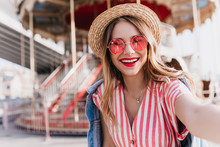 Pleased White Girl In Round Glasses Making Selfie Near Carousel. Outdoor Shot Of Cheerful Blonde Woman In Straw Hat Taking Picture Of Herself In Amusement Park.