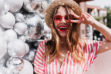 Wall Mural - Ecstatic european girl laughing while posing beside sparkle balls. Outdoor portrait of beautiful good-humoured lady in straw summer hat.