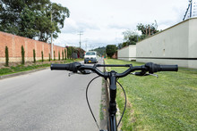 Close Up To A Black Mountain Bicycle Handle Bar With Grips Near To A Green Garden And Blurred White Van Transit Over Asphalt Way.
