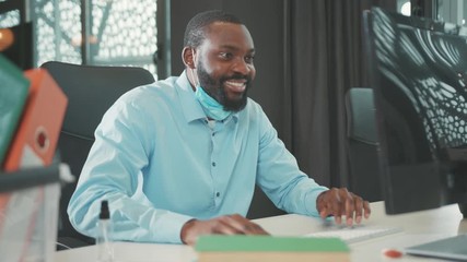 Happy smiling African American man wearing mask uses computer for work in modern office programmer looks at monitor screen businessman coronavirus close up slow motion