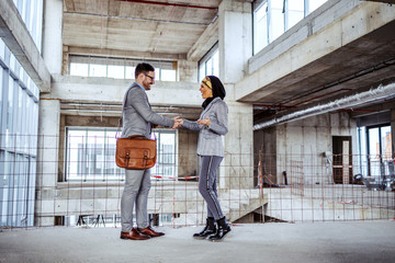 Wall Mural - Attractive muslim female investor meeting a sales agent and shaking hands with him while standing in future modern business center. Building in construction process interior.