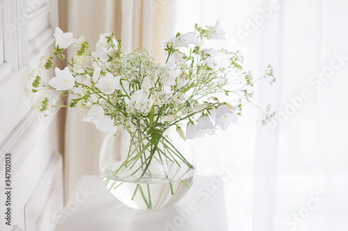Bouquet of gentle bells in vase. Morning light in the room. Soft home decor, glass vase with white flowers on  white wall background and on wooden table. Interior. Greeting card. Copy space.