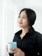 © nuiiko - Asian business woman drinking coffee near window in the morning, lifestyle concept.