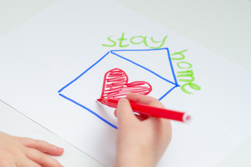 Wall Mural - Close up of hand of child drawing house with red heart on the sheet of paper with a painted words Stay Home over the house at home. Stay Home concept. Selective focus.