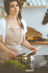 Wall Mural - Young brunette woman cooking soup in kitchen. Housewife holding wooden spoon in her hand. Food and health concept