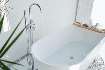 White bathroom modern interior. Luxurious decor with plants, window, spa at home. Bathtub is filled with tap water, nobody