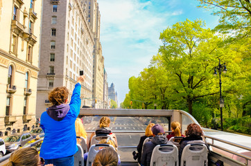 tourists enjoying views of new york city from top of open roof bus in spring; woman in blue is takin