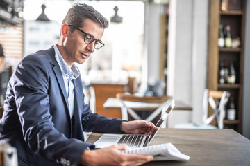 Young businessman in a jacket and glasses reads in a cafeteria with a computer on a table in front of him