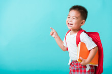 Back To School Concept. Portrait Asian Happy Funny Cute Little Child Boy Smile Hug Books And Point Finger To Side Away Space, Isolated Blue Background. Kid From Preschool Kindergarten With School Bag