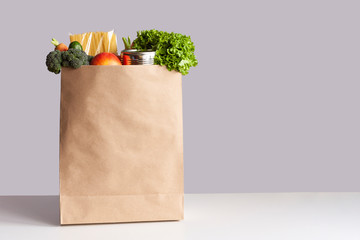 Various grocery items in paper bag on white table opposite gray wall. Bag of food with fresh vegetables, fruits, pasta and canned goods. Food delivery, shopping or donation concept. Copy space.