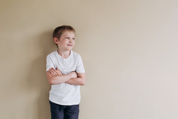 A happy boy of European appearance in a white T-shirt looks into the distance. Photo of a 7 years old baby on a beige background.