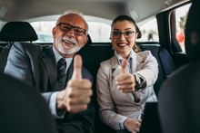 Good Looking Senior Business Man And His Young Woman Colleague Or Coworker Sitting On Backseat In Luxury Car. They Are Smiling And Showing Thumb Up. Transportation In Corporate Business Concept.