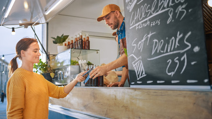 Food Truck Employee Hands Out a Burger to a Happy Young Woman. Young Lady is Using NFC Payment on Her Smartphone to Pay for Food. Street Food Truck Selling Burgers in a Modern Hip Neighbourhood.