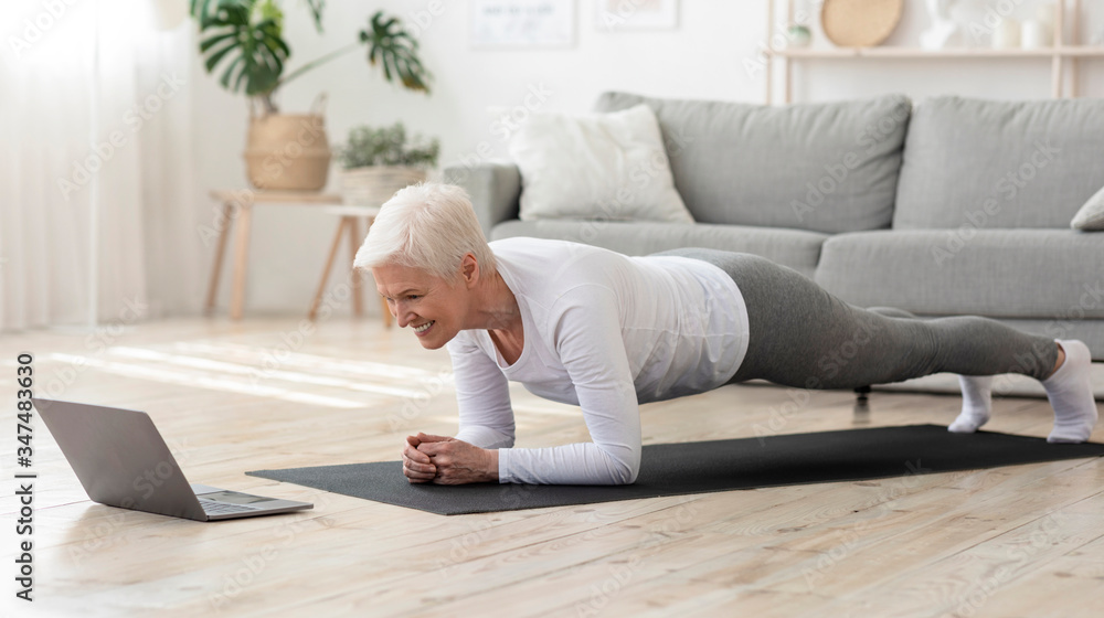 Sporty senior woman doing yoga plank while watching tutorial on laptop - obrazy, fototapety, plakaty 
