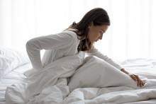 Side View Of Millennial Woman Woke Up Sitting In Bed, Unhealthy Female Woke Up Feels Severe Lower Back Pain During Period, Pinched Nerve Discomfort In Vertebrae, Herniated Spinal Disc Symptoms Concept