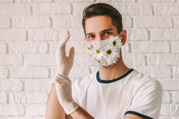 Portrait of happy male doctor in daisy flower medical face mask put on latex protective gloves. Young man in summer protective face mask put on sterile medical gloves for COVID-19 personal protection