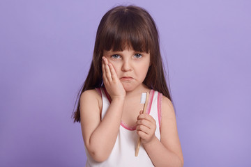 Wall Mural - Horizontal portrait of poor sad child having disappointed facial expression, being sad, having toothache, holding toothbrush, looking directly at camera, wearing sleep shirt. Children concept.
