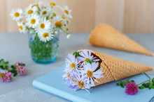 Bouquet Of Chamomiles In A Waffle Cone On A Gray Background And Blue Book. The Flowers Are Blooming. Wildflowers.