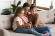 © fizkes - Smiling mother with two little daughters using phone together, sitting on cozy sofa in living room, cute preschool girls with mum watching video on smartphone, playing game or shopping online