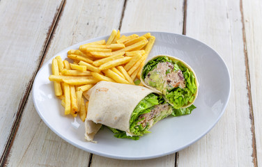 Wall Mural - Pita bread pancake stuffed with meat, lettuce, red pepper and fried potatoes on a white plate, on a wooden table. Close-up.