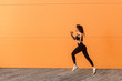 © khosrork - Motivated confident fit woman athlete in tight sportswear, black pants and top, starting to run, jogging outdoor against orange wall, advertising area. Health care and weight loss, sport activity