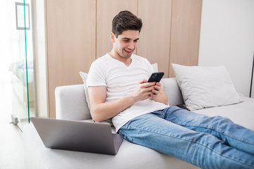 Young handsome man sitting relaxed on sofa with laptop checking his smartphone messages