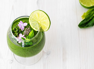 Spinach smoothie with fruits and seeds superfoods, decorated with mint leaves, a slice of lime and wild flowers in a glass cup. Top view, white wooden background