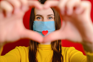 Cute girl making heart shape hand gesture looking at camera, wear medical mask with a red heart on it as a way to show appreciation and to thank all essential employees during the Covid-19 pandemic