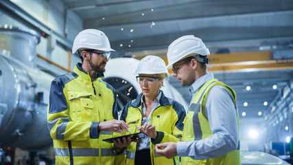 Wall Mural - Three Heavy Industry Engineers Stand in Pipe Manufacturing Factory, Use Digital Tablet Computer, Have Discussion. Large Pipe Assembly. Design and Construction of Oil, Gas and Fuels Transport Pipeline