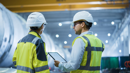 Wall Mural - Two Heavy Industry Engineers Stand in Pipe Manufacturing Factory, Use Digital Tablet Computer, Have Discussion. Construction of Oil, Gas and Fuels Transport Pipeline. Back View Sparks Flying
