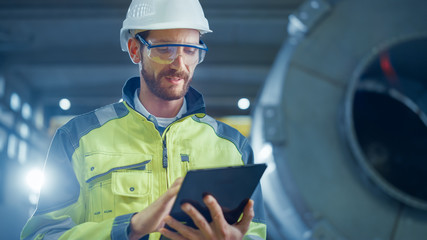 Wall Mural - Portrait of Professional Heavy Industry Engineer / Worker Wearing Safety Uniform and Hard Hat Uses Tablet Computer. In the Background Construction Factory for Oil, Gas and Fuels Transport Pipeline