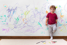 Child Mischief. Boy With A Distracted Face Because He Drew The Entire Wall. Little Boy Leaning Against The White Wall Where He Made Many Drawings With Colored Pencils. Kid Indoors, At Home.