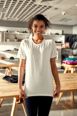 Wall Mural - You would like it. Young woman smiling at camera, while trying on t shirt, standing in the store. Clothes at custom T-shirt, clothing printing company