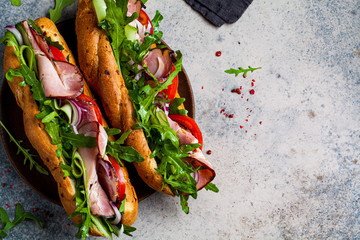 Two fresh baguette sandwiches with meat, tomato, cucumber and arugula on gray background, top view.