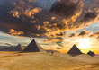 A dramatic sunrise with impressive clouds over the desert and pyramids at Giza in Egypt. A man in a turban is riding a camel. The sun casts long shadows.