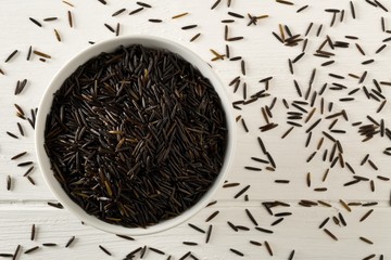 Wall Mural - Heap of uncooked, raw, black wild rice grains in white bowl on white wooden table background, flat lay top view from above