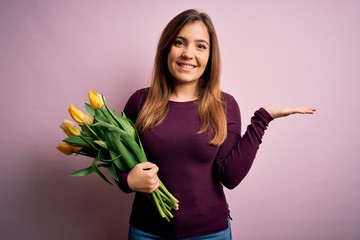 Sticker - Young blonde woman holding romantic bouquet of yellow tulips flowers over pink background smiling cheerful presenting and pointing with palm of hand looking at the camera.