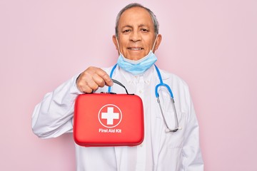 Senior handsome hoary doctor man wearing stethoscope and medical mask holding first aid kit with a happy face standing and smiling with a confident smile showing teeth