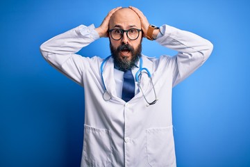 Canvas Print - Handsome bald doctor man with beard wearing glasses and stethoscope over blue background Crazy and scared with hands on head, afraid and surprised of shock with open mouth