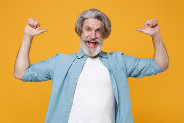 Excited elderly gray-haired mustache bearded man in casual blue shirt posing isolated on yellow background studio portrait. People lifestyle concept. Mock up copy space. Pointing thumbs on himself.