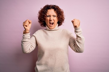 Poster - Middle age beautiful curly hair woman wearing casual turtleneck sweater over pink background angry and mad raising fists frustrated and furious while shouting with anger. Rage and aggressive concept.