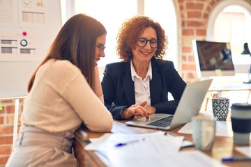 Canvas Print - Two beautiful businesswomen smiling happy and confident. Sitting with smile on face working together using laptop at the office