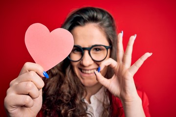 Sticker - Young beautiful woman with curly hair holding paper heart over isolated red background doing ok sign with fingers, excellent symbol