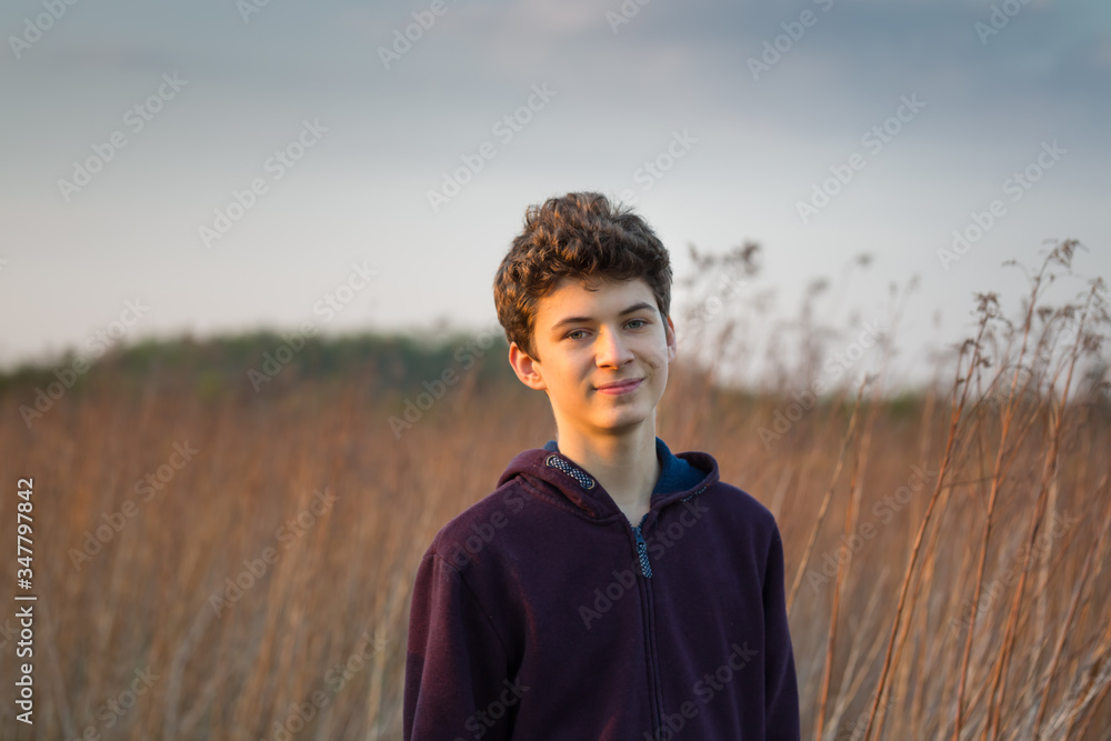 delikatnie uśmiechnięty chłopak nastolatek w bluzie z kapturem na polu wysokich traw, oświetlony  ciepłym światłem zachodu słońca - obrazy, fototapety, plakaty 