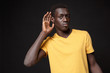 © ViDi Studio - Confused young african american man guy in yellow t-shirt posing isolated on black background studio portrait. People lifestyle concept. Mock up copy space. Try to hear you with hand gesture near ear.