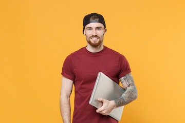 Smiling young bearded tattooed man guy in casual t-shirt black cap posing isolated on yellow background studio portrait. People emotions lifestyle concept. Mock up copy space. Hold laptop pc computer.