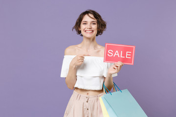 Smiling young woman in summer clothes isolated on violet background. Shopping discount sale concept. Mock up copy space. Hold package bag with purchases pointing index finger on sign with SALE title.